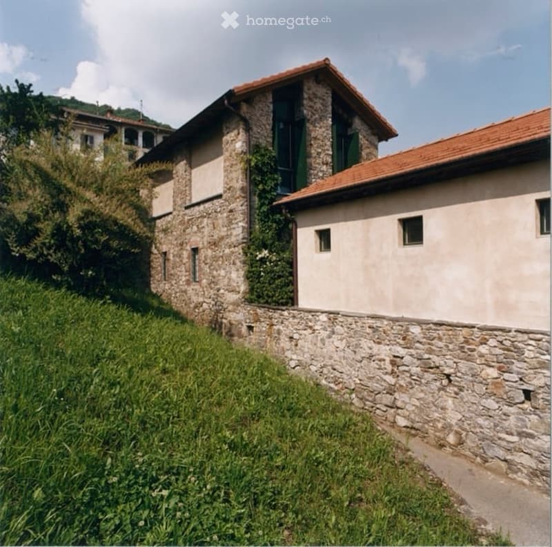 Two-story stone building with a raised ground floor, red-tiled roof, and green shutters on the upper level. The lower level has a stone wall and a small grassy area.