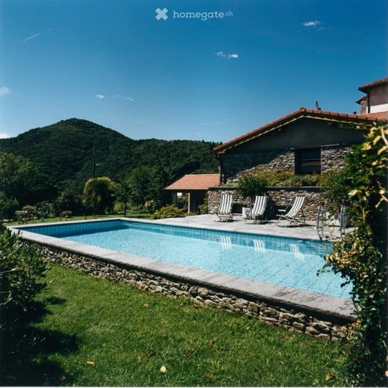A rectangular swimming pool surrounded by a stone border, with lounge chairs and a small table nearby. The pool is set in a grassy area with a stone house in the background and a scenic mountain view.