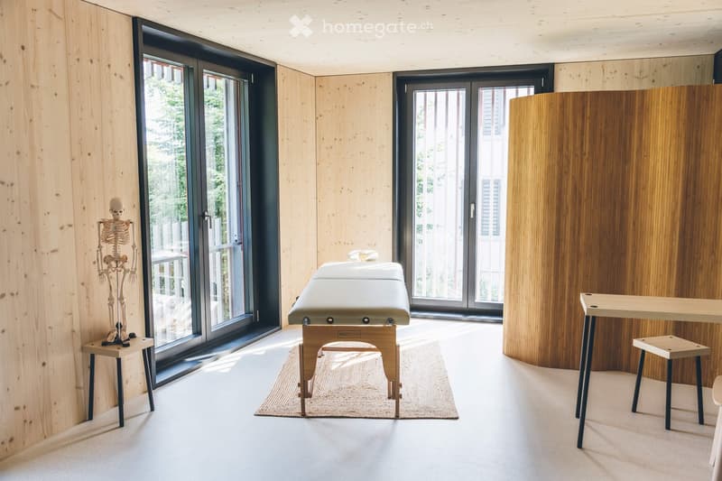 Modern room with light wooden walls, large glass doors, massage table, skeleton model, and small wooden stools.