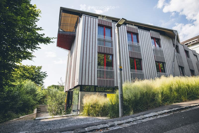 Modern two-story building with white and red exterior, large windows, balconies, and surrounding greenery.
