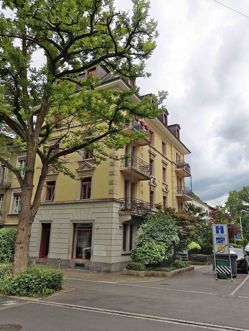 A multi-story residential building with yellow facade, red-framed windows, and balconies. The building is surrounded by greenery and trees, with a paved street in front.