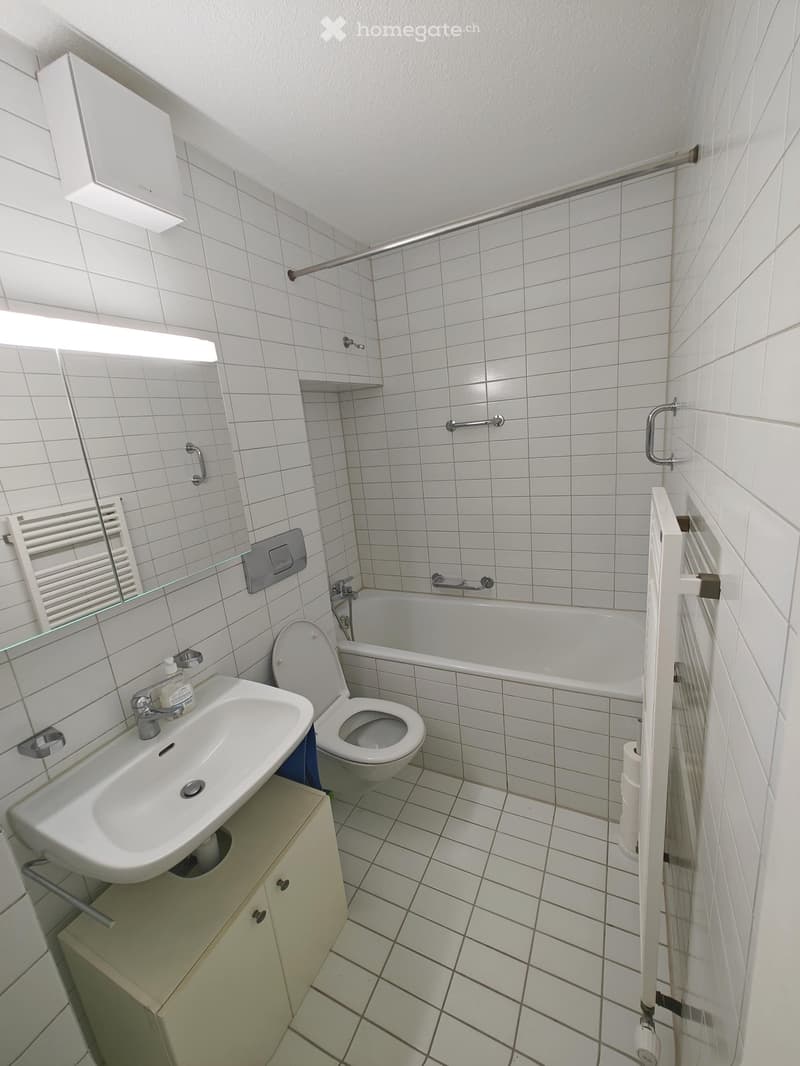 A small bathroom with white tiled walls and floor, featuring a sink, toilet, bathtub, and a mirror above the sink. A towel rack is mounted on the wall, and a small cabinet is placed under the sink.
