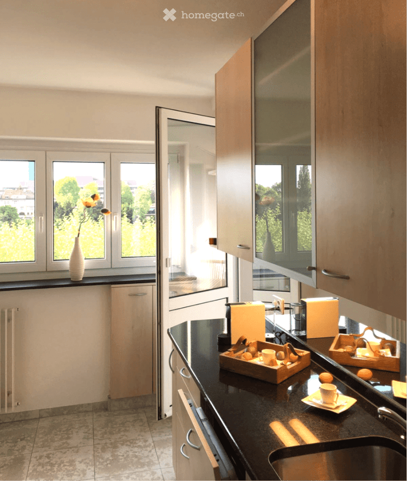Modern kitchen with black countertops, wooden cabinets, and a view of greenery through large windows. Includes a sink, faucet, cup, vase with flowers, and decorative trays.