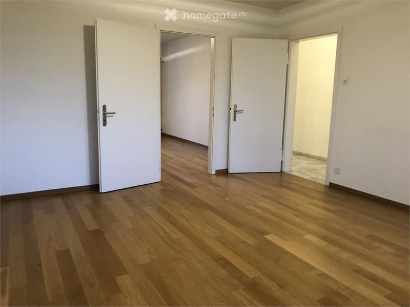 Empty room with wooden flooring and white walls, featuring multiple open doors leading to adjacent spaces.