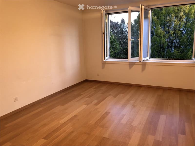 Empty room with wooden flooring and large window with open shutters, showing greenery outside.