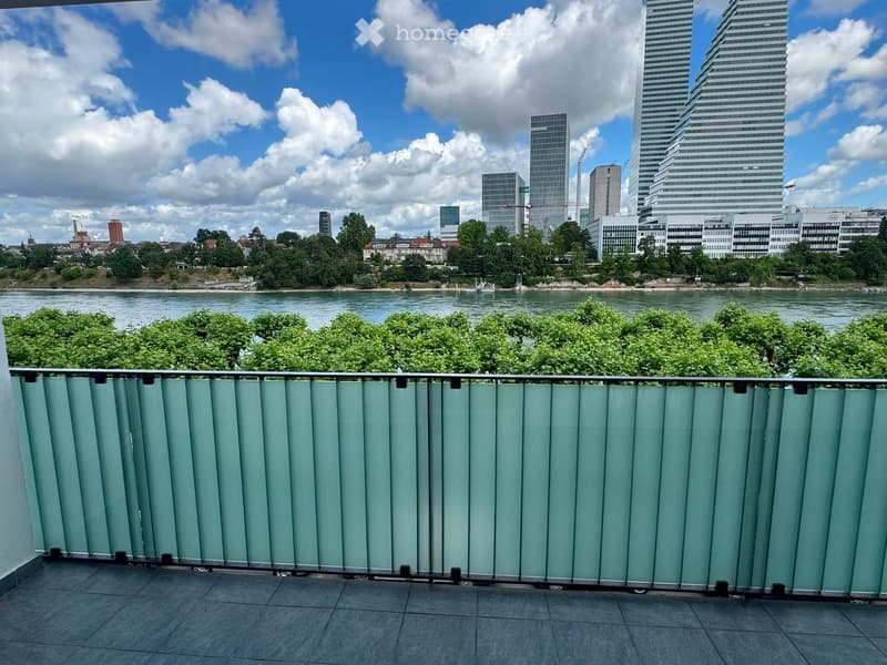 Balcony with modern glass railing, overlooking a river and cityscape with tall buildings and trees.