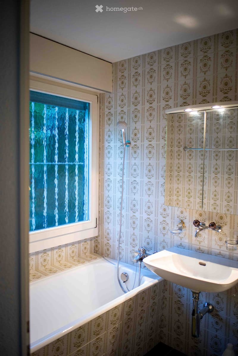Bathroom with a bathtub, sink, and showerhead. The walls are tiled with a decorative pattern, and there is a window with frosted glass.