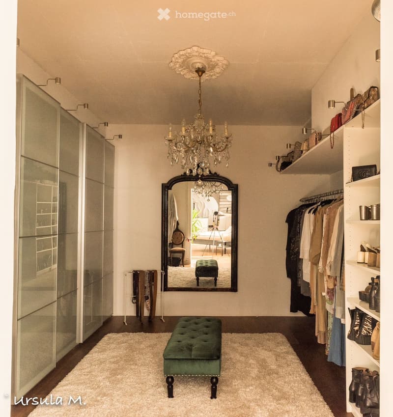 A well-lit, stylish room with a green tufted stool, a large ornate mirror, a wardrobe with hanging clothes, shelves with various items, and a chandelier hanging from the ceiling.