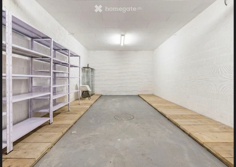 Empty room with white brick walls, concrete floor, metal shelving unit, and a small glass display case with a chair nearby.