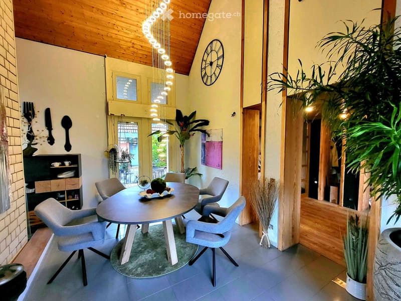 Modern dining area with a round table, four chairs, a hanging light fixture, and a potted plant. The room features wooden beams on the ceiling and a view of the outdoor area through large windows.
