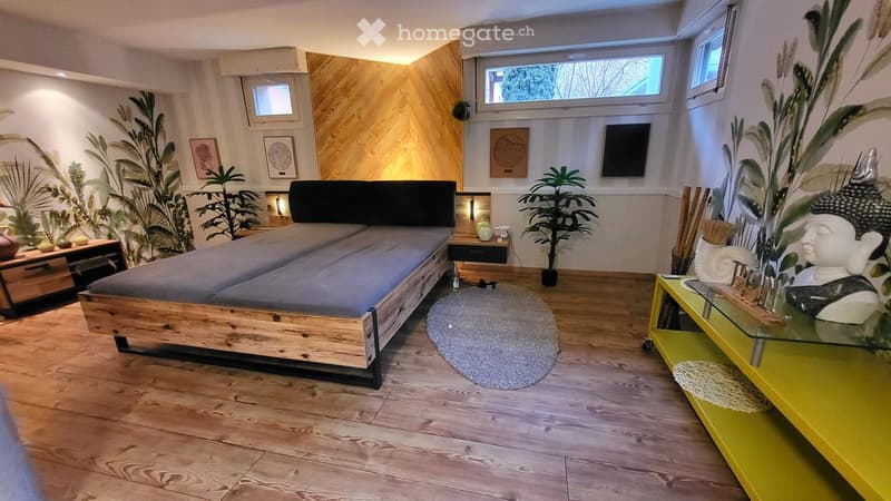 Modern, minimalist bedroom with wooden bed frame, grey bedding, nightstand, small table, and decorative plants.