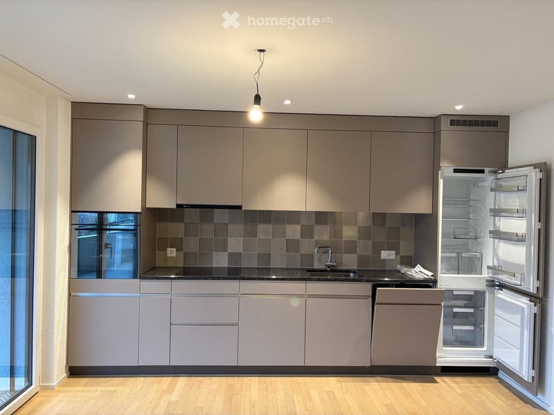 Modern kitchen with light grey cabinets, dark countertops, and stainless steel appliances including a refrigerator and dishwasher. Features a large window and pendant lighting.