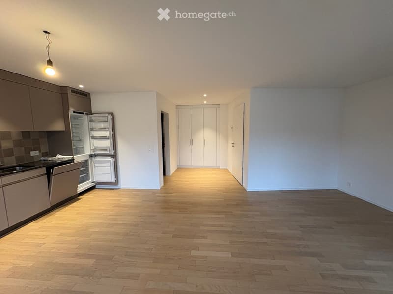 Modern kitchen with light wooden flooring, grey cabinets, and stainless steel appliances including a refrigerator and oven. The space is open and well-lit with recessed lighting.