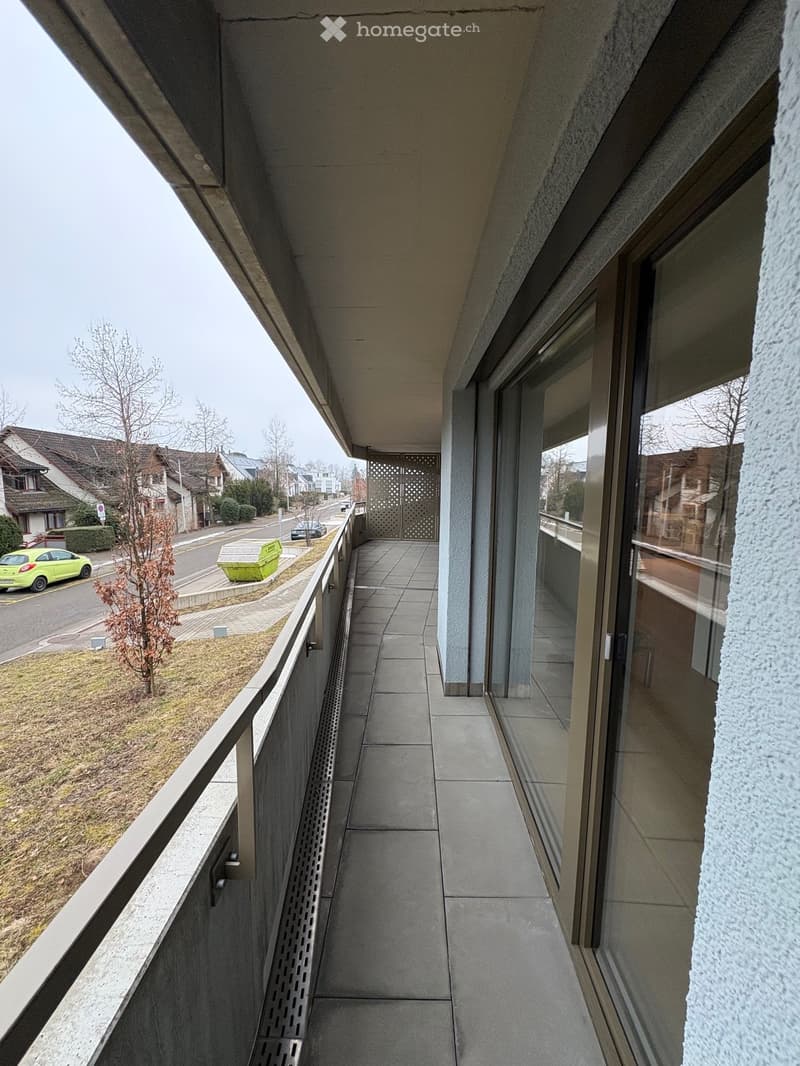 Modern balcony with tiled floor, metal railing, and large glass windows overlooking a residential street.