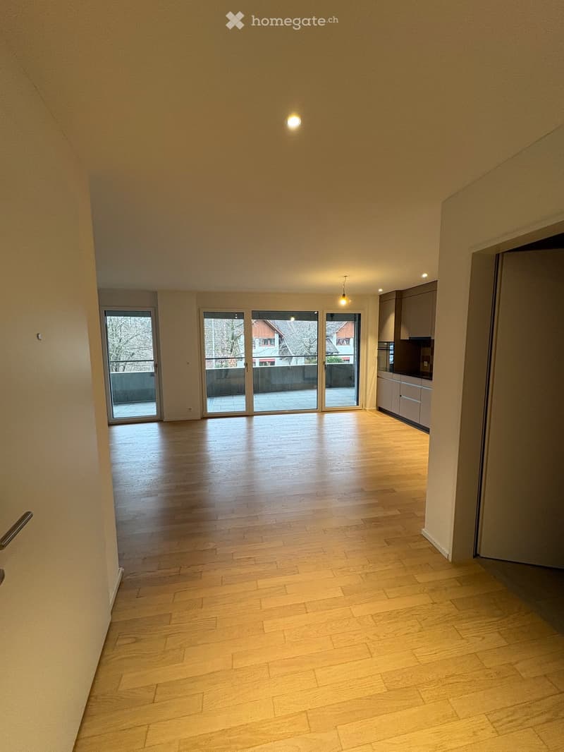 Empty room with wooden flooring, large sliding glass doors leading to a balcony, and a small kitchen area visible on the right side.