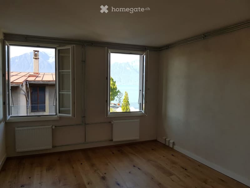 Empty room with wooden flooring, large windows offering a scenic mountain and lake view, radiators, and basic electrical outlets.
