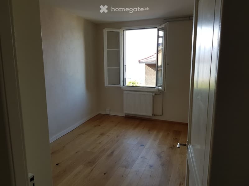 Empty room with wooden flooring, white walls, and a radiator under a large window that is open, allowing natural light to enter.