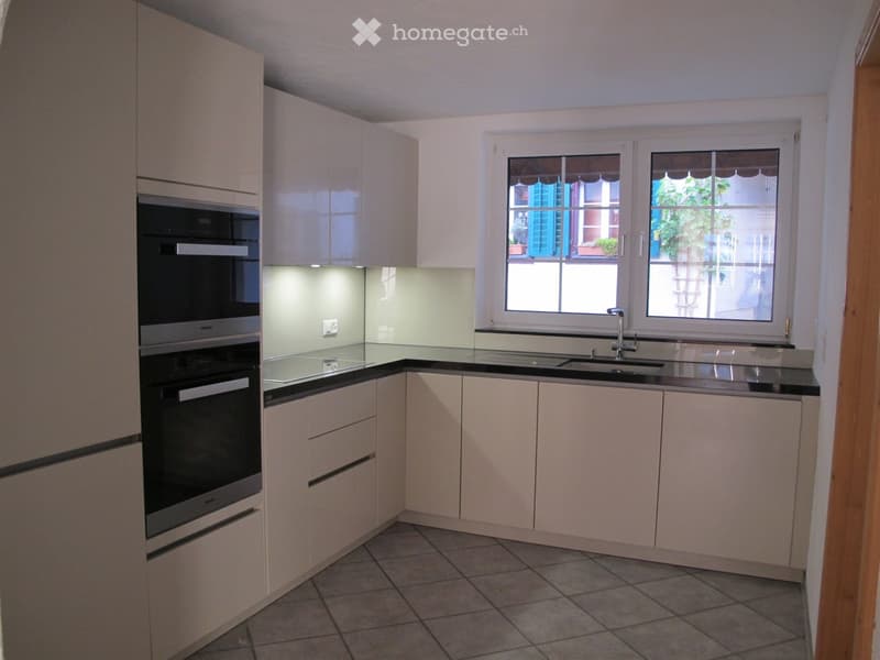 Modern kitchen with light-colored cabinets, black countertops, and a built-in oven. Features a double sink with a modern faucet and large windows offering a view of the outside.