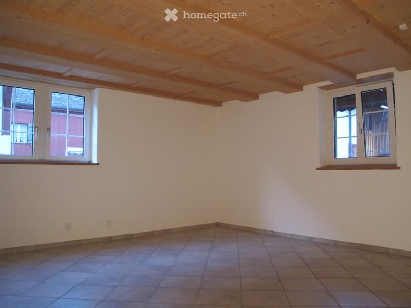 Empty room with tiled floor, exposed wooden ceiling beams, and large windows with white frames. The walls are painted a light beige color, and there are small built-in wooden shelves above the windows.