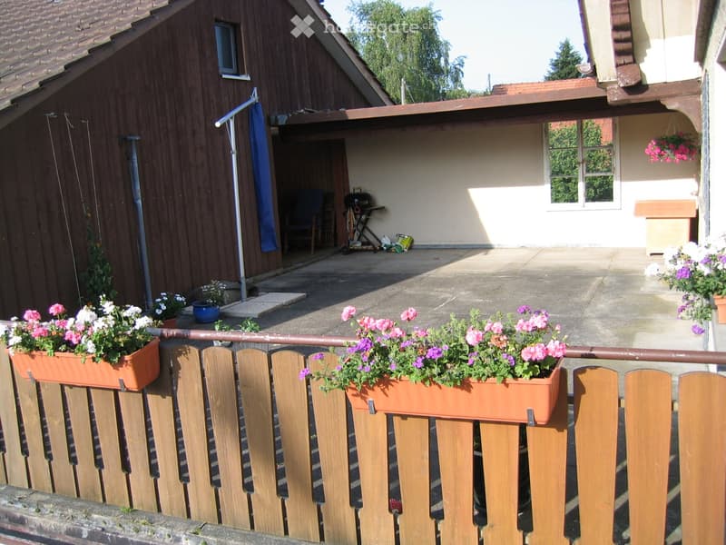 A wooden fence with flower boxes, a patio area with a grill, a blue umbrella, and a small wooden shed.