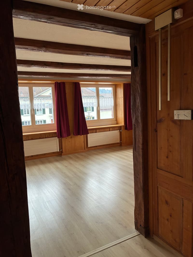 Empty room with wooden beams on the ceiling, wooden floor, large windows with red curtains, radiator, and a clock on the wall.