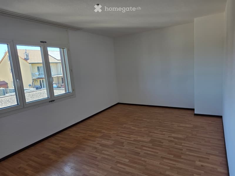 Empty room with wooden flooring and large windows, showing a view of the street and neighboring buildings.
