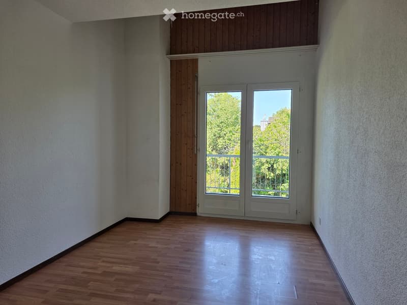 Empty room with wooden flooring, white walls, and a large window with a view of greenery outside.