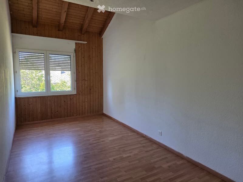 Empty room with wooden flooring, white walls, and a window with blinds. The ceiling features exposed wooden beams.