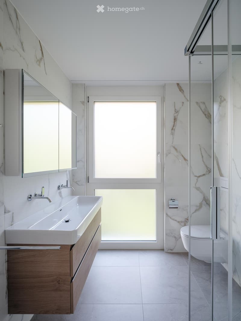 Modern bathroom with a white sink, wooden cabinet, large mirror, and a glass-enclosed shower area with marble accents.