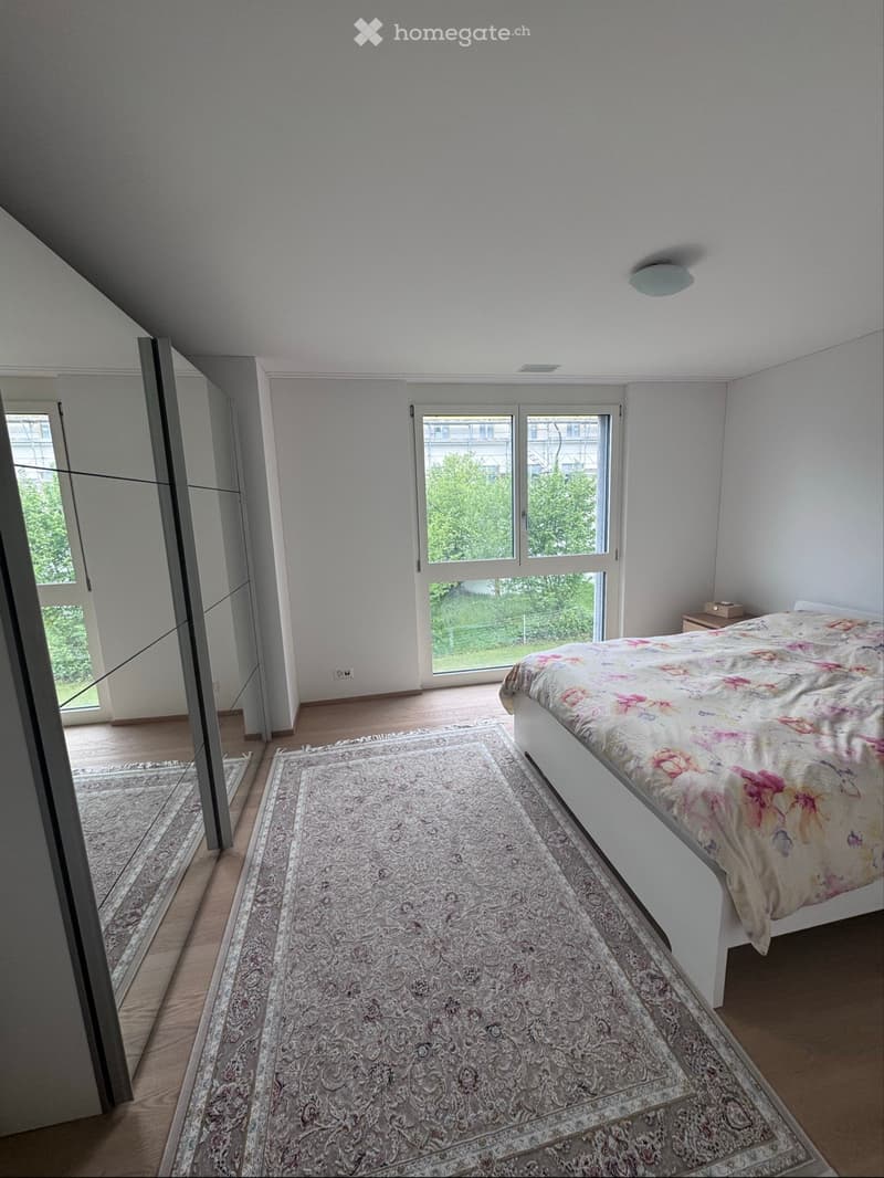 Bright bedroom with wooden flooring, large window, bed with floral bedding, and patterned rug.