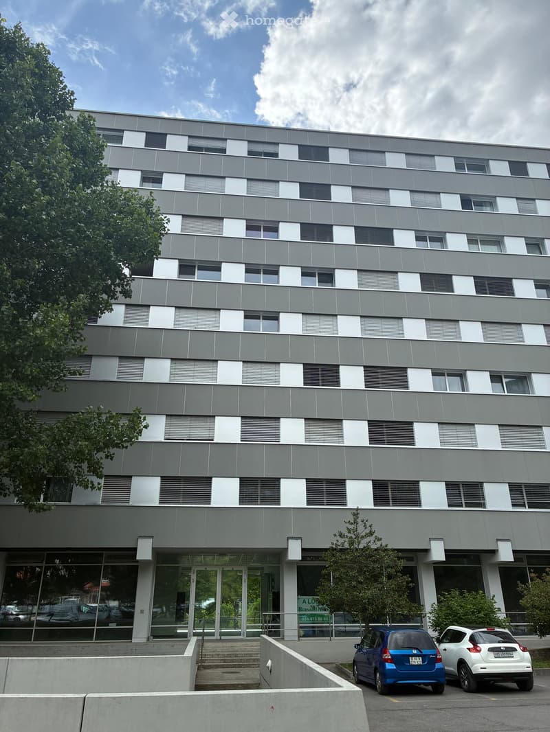 Modern multi-story building with grey and white facade, large windows, and a glass entrance. Two cars are parked in front.