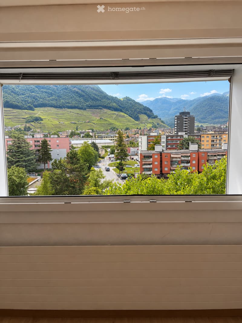 Empty room with a large window offering a scenic view of a town, green hills, and mountains. The room has light-colored walls and wooden flooring.