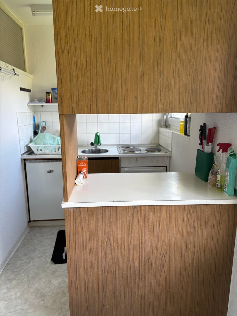 Modern kitchen with wooden cabinets, white countertop, sink, stove, oven, and dishwasher. Cleaning supplies and a knife holder are visible on the counter.
