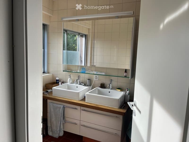 Modern bathroom with double sinks, a large mirror, and a wooden countertop. The space includes a towel, toiletries, and a cabinet below the sinks.