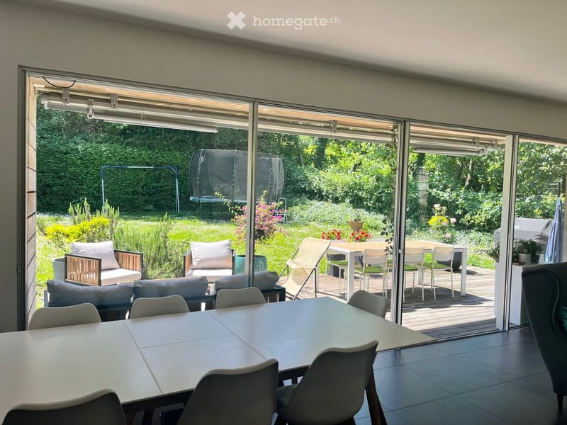 Bright room with large sliding glass doors opening to a garden, featuring a dining table with chairs, a sofa with pillows, and an ottoman.