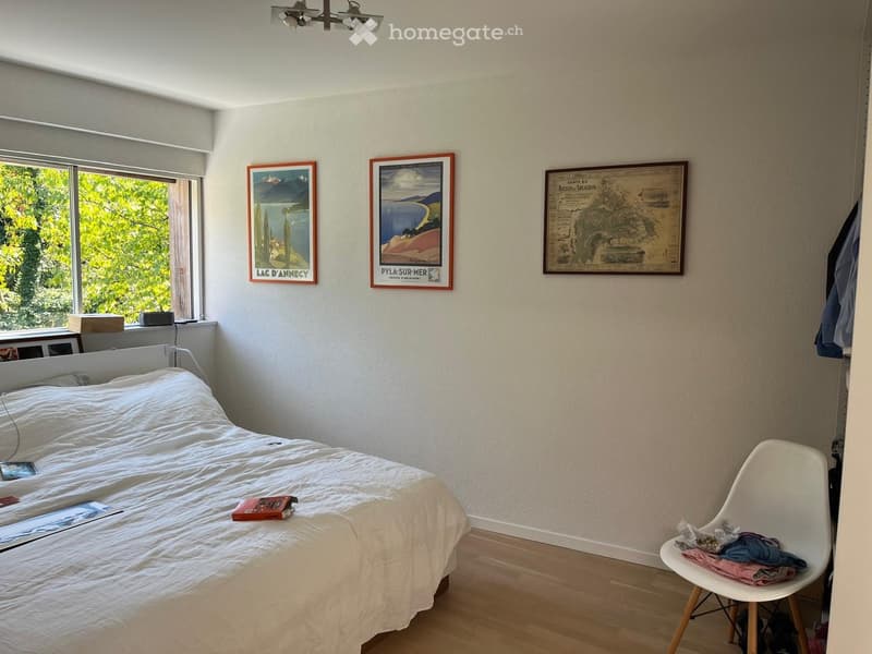 A well-lit bedroom with a white bed, a modern white chair, and three framed pictures on the wall. The room has wooden flooring and large windows with a view of greenery.