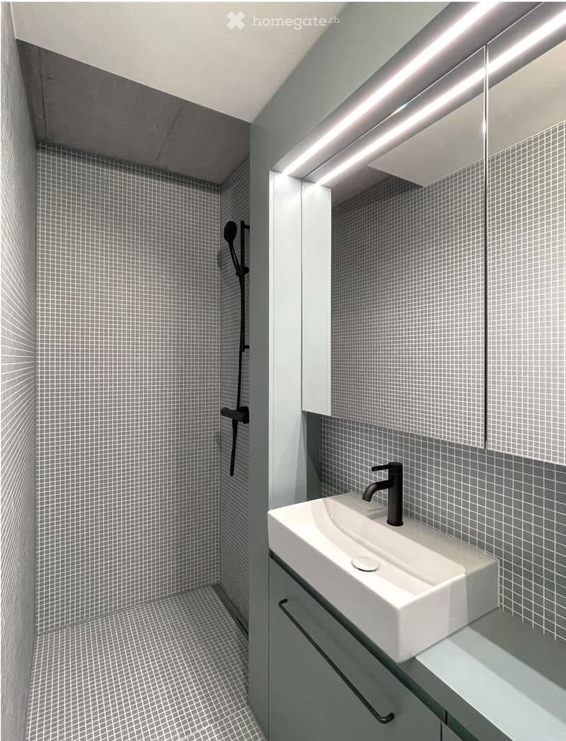 Modern bathroom with grey tiled walls and floor, white sink, black faucet, and built-in mirror with LED lighting.