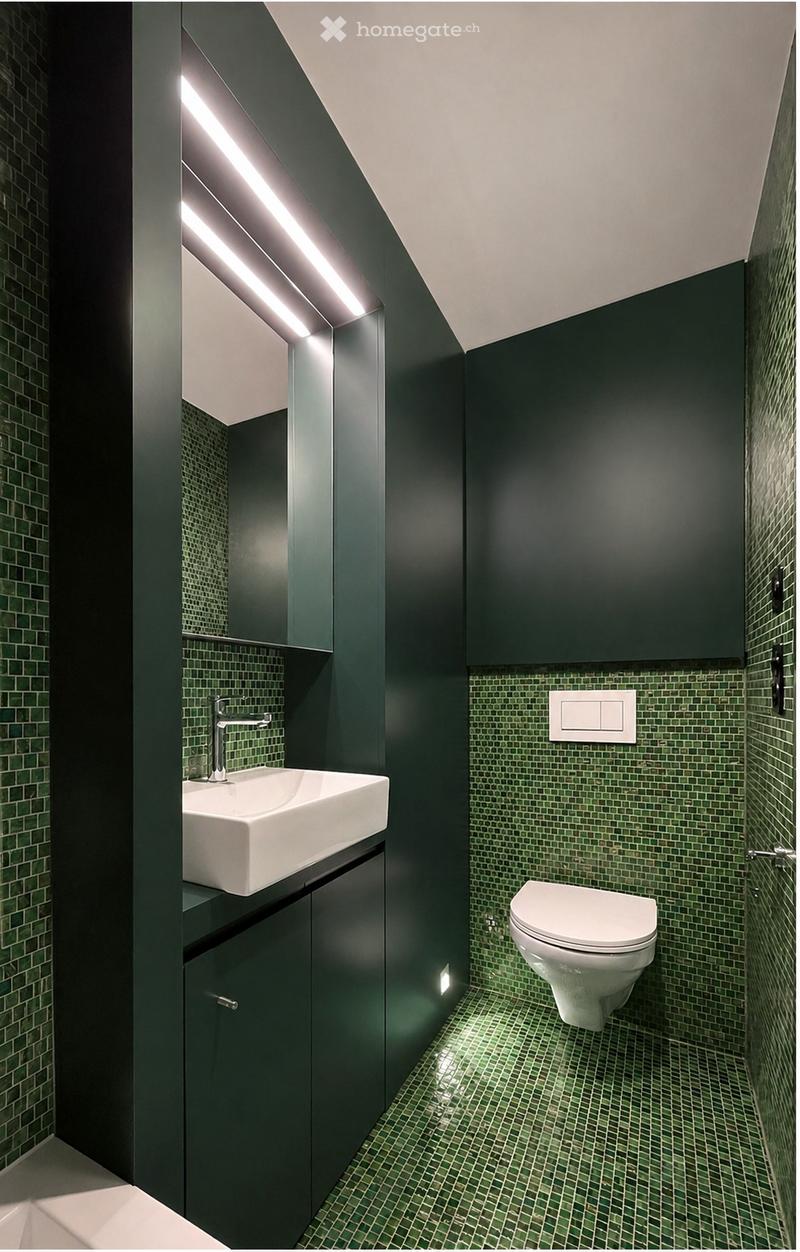Modern bathroom with dark green tiles, white sink, chrome faucet, and toilet. Features built-in dark green cabinets and recessed lighting.