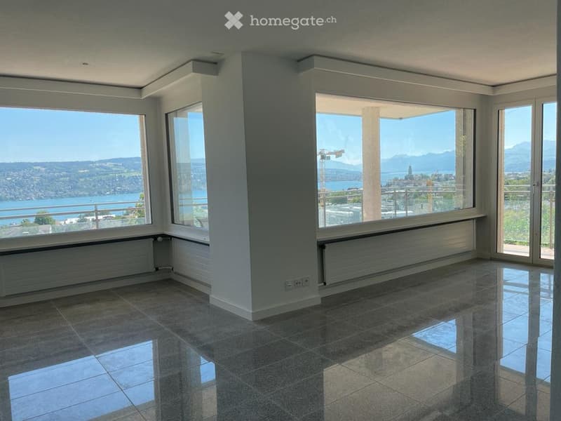 Bright, unfurnished room with large windows offering a scenic view of a lake and mountains. The room features polished tile flooring and modern white walls.