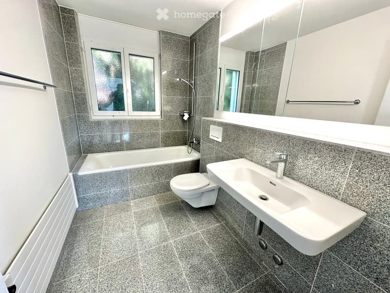 Modern bathroom with grey tiled walls and floor, featuring a bathtub, toilet, sink, and large mirror.