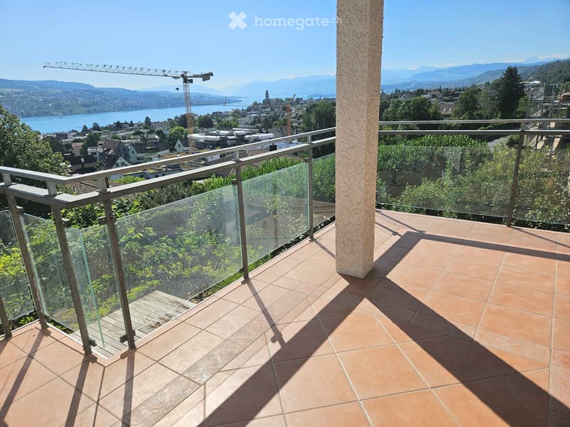 Balcony with glass railing, tiled floor, scenic view of city, lake, and mountains.