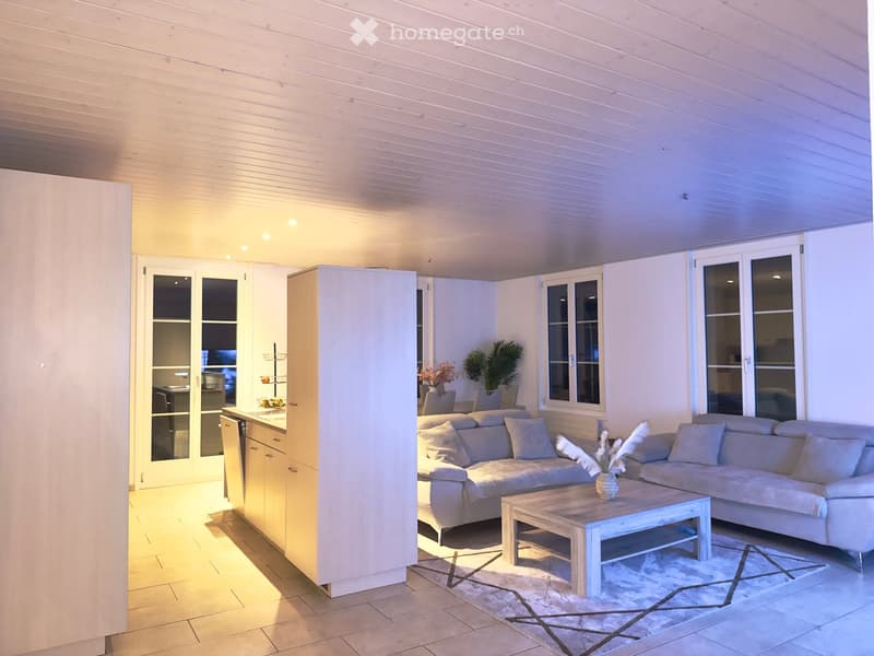 Bright living room with a light-colored sofa, coffee table, and a small kitchen area with cabinets and a refrigerator. The space features large windows and a modern, minimalist design.