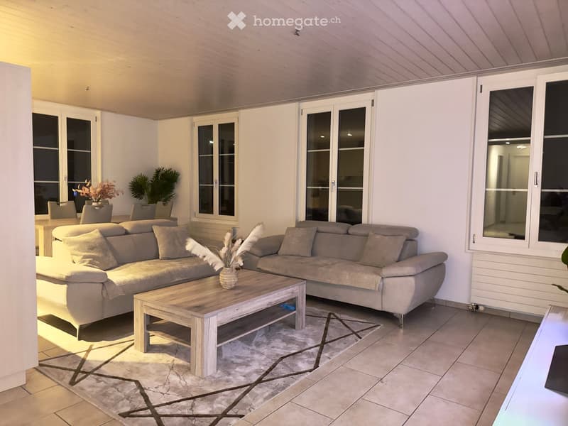 A modern, well-lit living room with a large sectional sofa, a wooden coffee table, and a few chairs. The room features large windows, a tiled floor, and decorative plants.