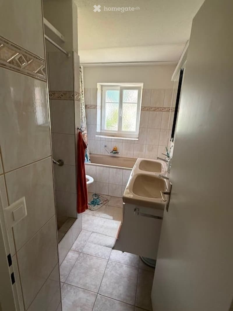 Bathroom with a double sink, bathtub, toilet, and a window above the bathtub. The walls are tiled, and there is a shower curtain visible.
