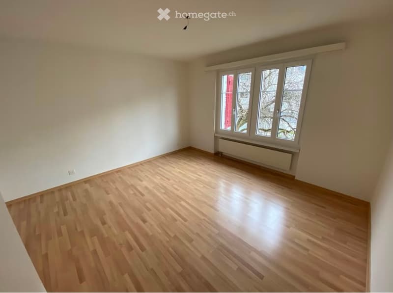 Empty room with light wooden flooring, white walls, and large windows with white frames. No furniture or equipment is present.