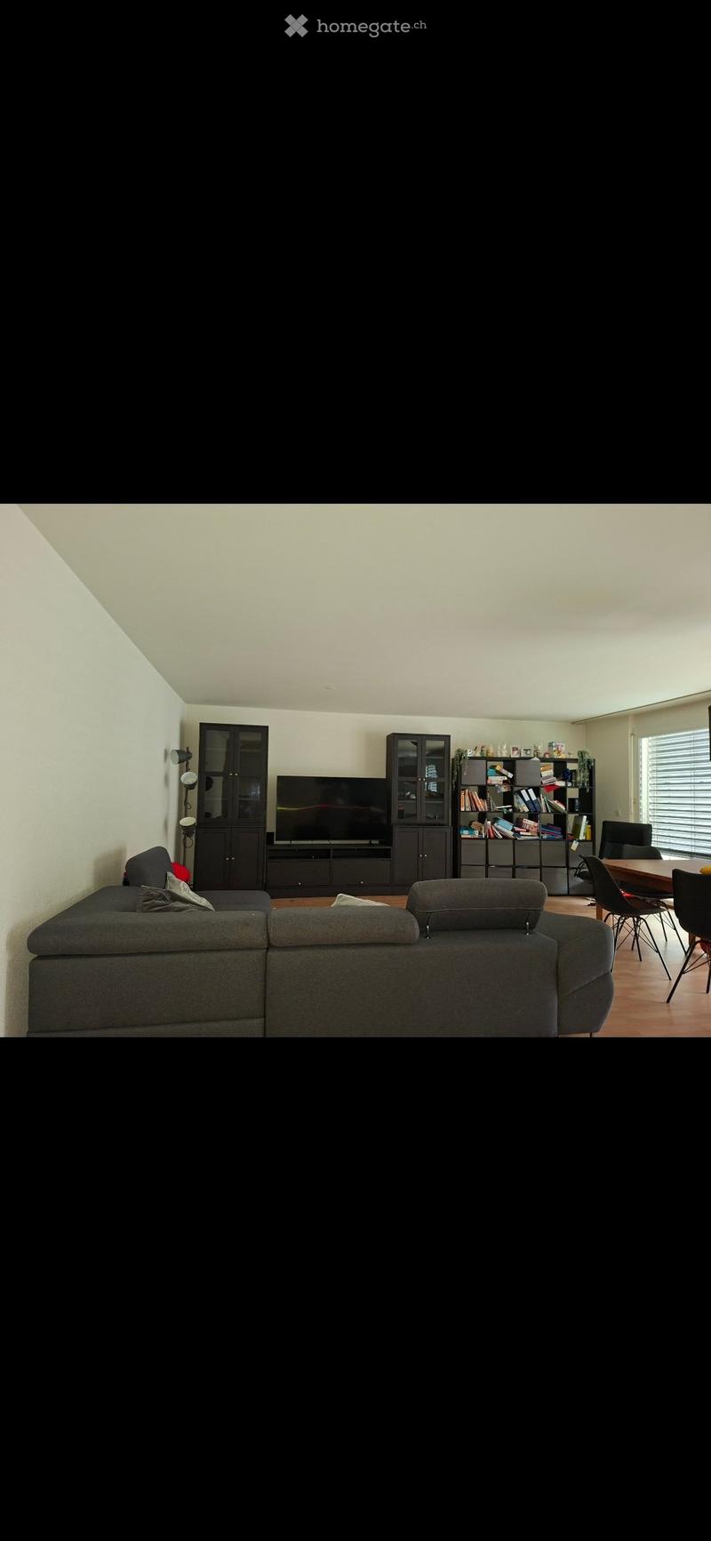 Modern living room with a grey sectional sofa, flat-screen TV, dark wooden cabinets, and a dining area with chairs and a table.