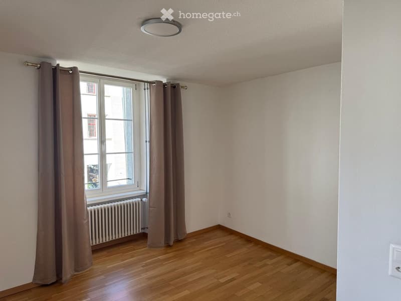 Empty room with wooden flooring, white walls, large window with brown curtains, radiator, and a ceiling light fixture.
