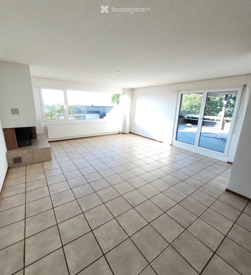 Empty room with tiled floor, large windows, sliding glass door leading to outdoor area, and a small built-in bench with a fireplace.