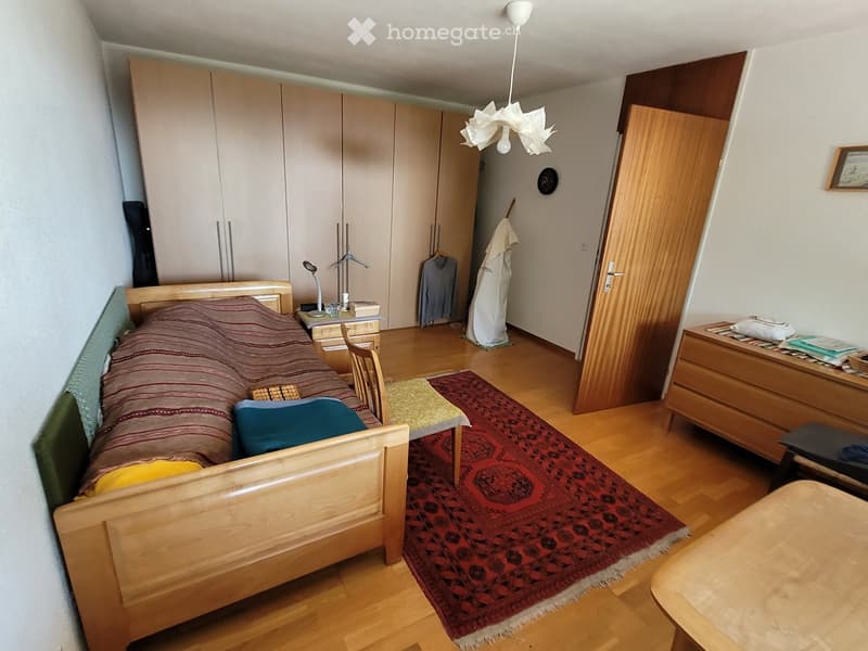 Cozy bedroom with wooden bed, nightstand, chair, small table, wardrobe, and mirror. Features a patterned rug and a hanging light fixture.