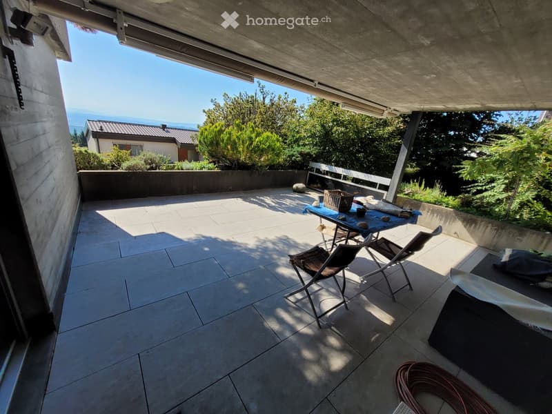 A modern, tiled balcony with a table and chairs, overlooking a garden and distant sea view.